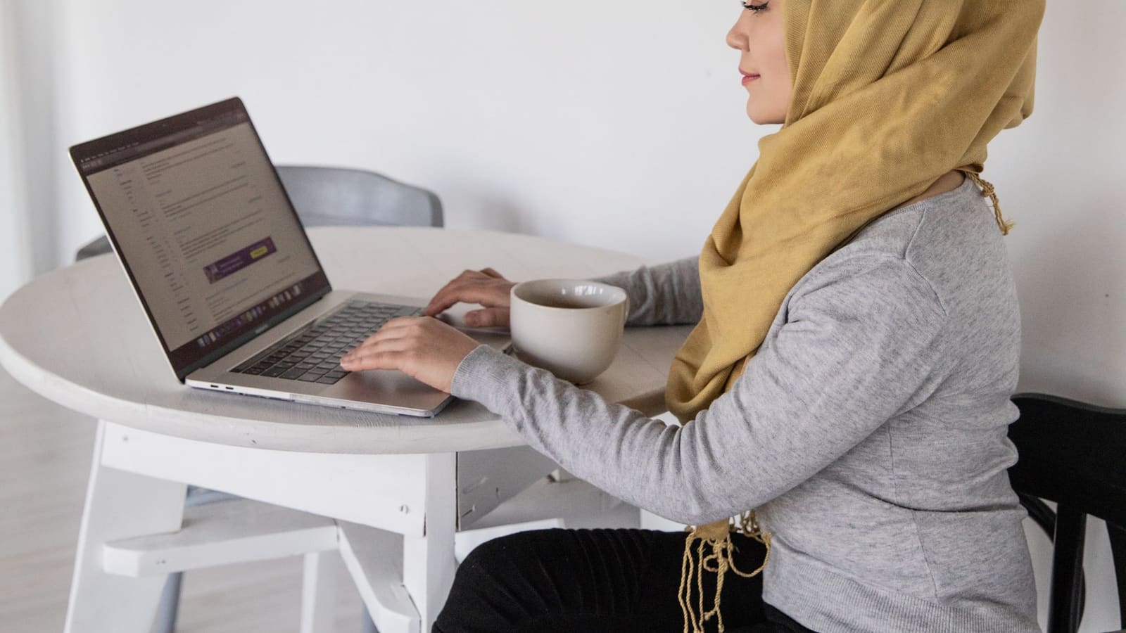 Perempuan berhijab bekerja di depan laptop sambil memegang cangkir