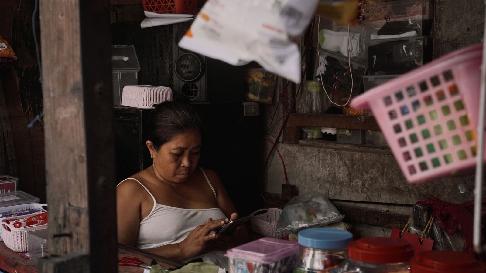 Pemilik warung kecil duduk di antara dagangannya sambil menatap layar ponsel